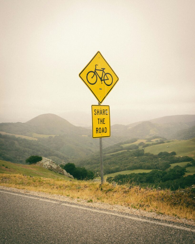 A scenic country road with a 'Share the Road' bike lane sign amidst rolling hills on a cloudy day.