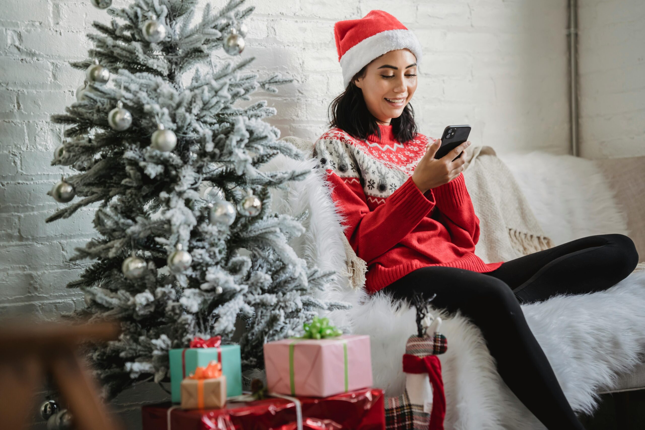 Smiling woman in Santa hat using smartphone by decorated tree with gifts.